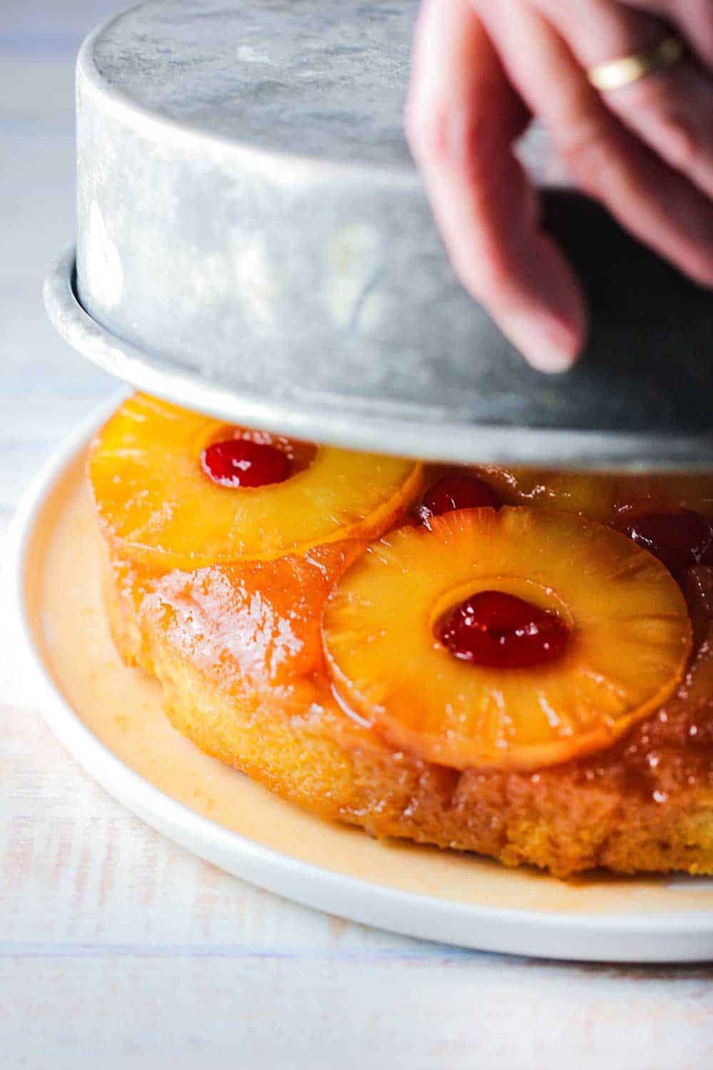 Two hands pulling an inverted cake pan away from a pineapple upside-down cake.