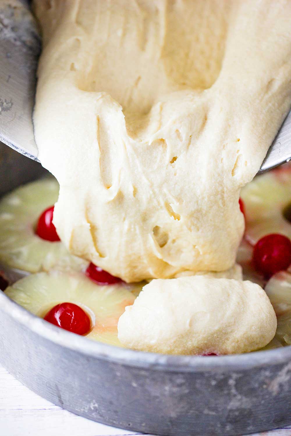 Yellow butter cake batter being poured into a cake pan with pineapples and cherries. 