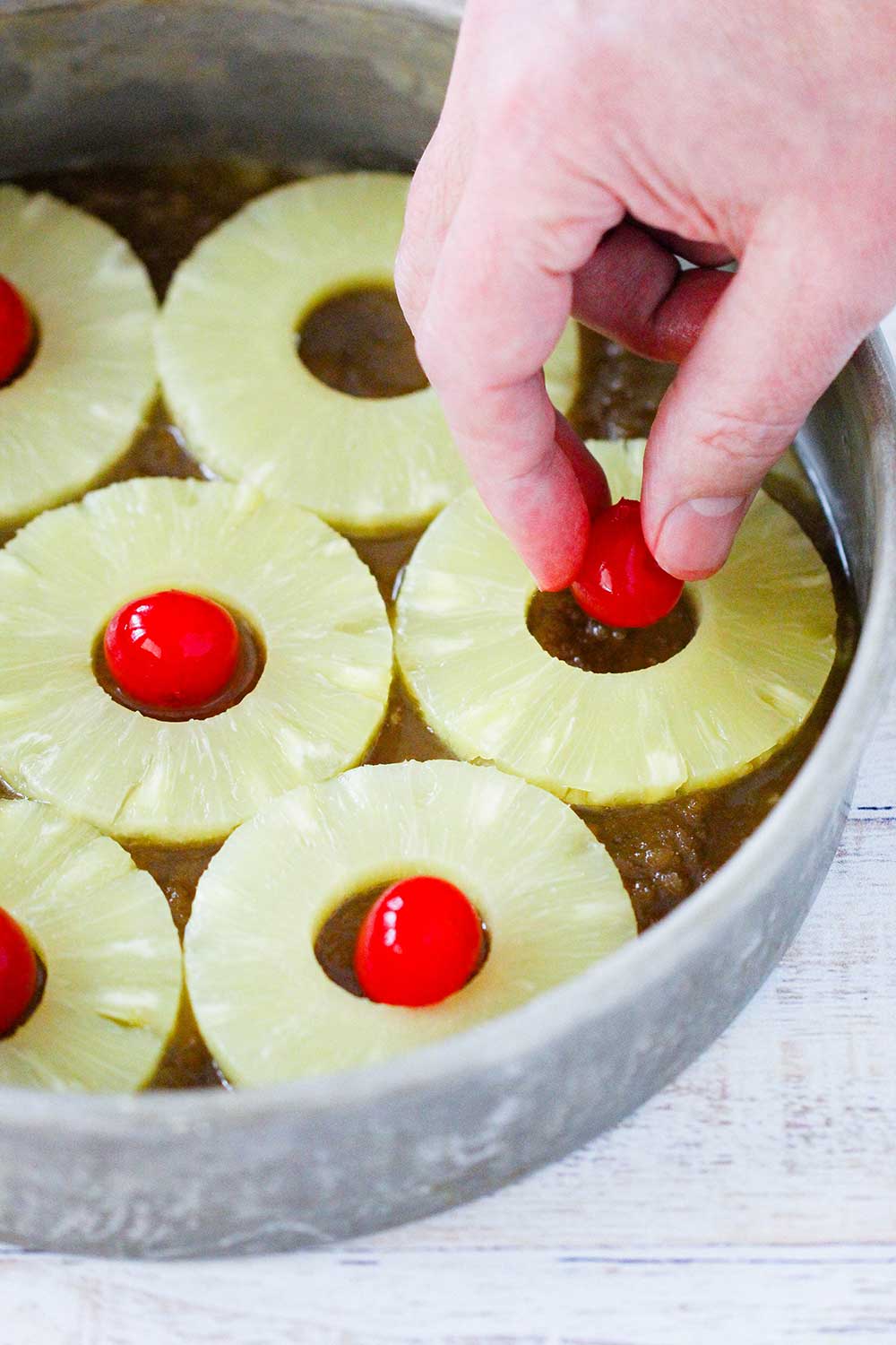 A hand placing a cherry in the whole of a pineapple ring in a cake pan. 