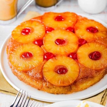 A pineapple upside-down cake on a white platter next to a glass of orange and a cup of coffee.