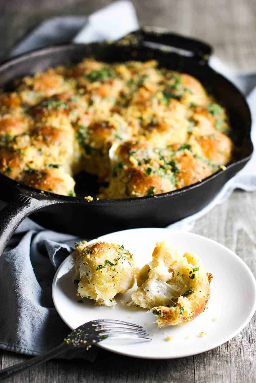 Cheesy skillet rolls in the background in a cast iron skillet with a single roll on a plate in front