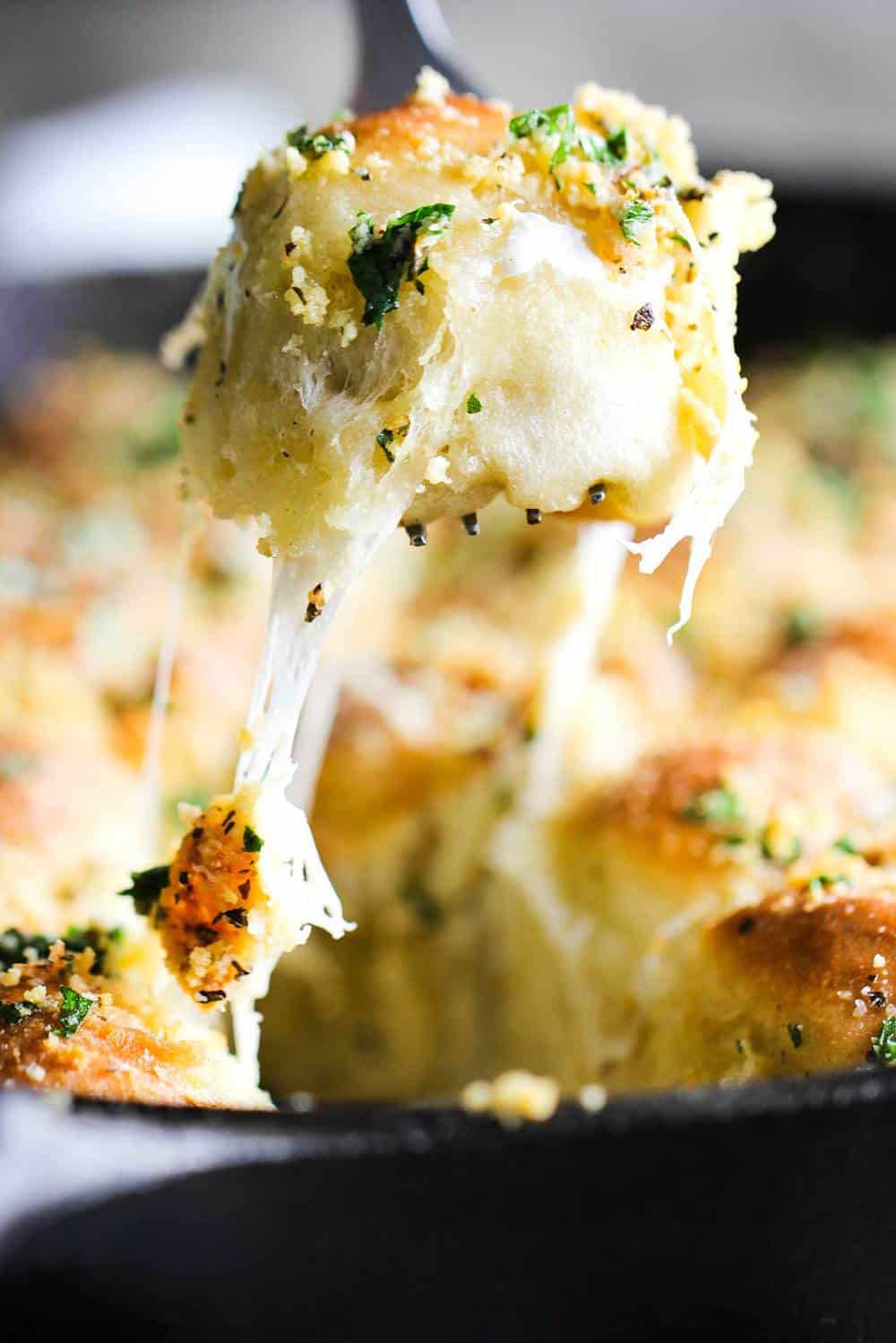 A cheesy skillet roll being pulled out of a cast iron skillet with gooey cheese and herbs on it. 