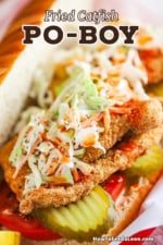 A close-up view of a fried catfish po-boy with homemade coleslaw and hot sauce sitting on top of the catfish, pickles, and sliced tomatoes, all on a soft roll.