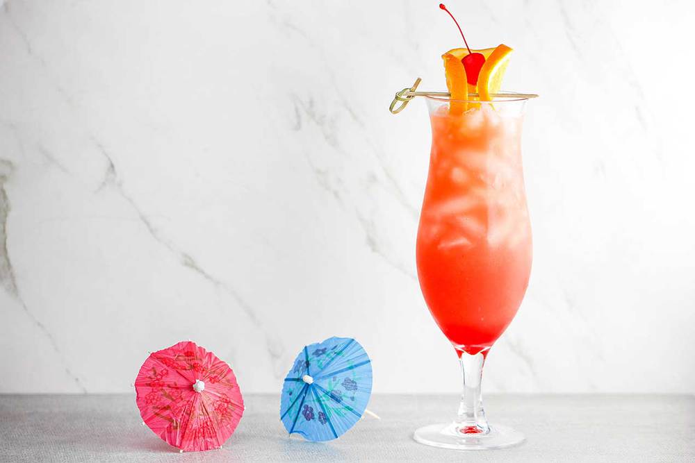 A hurricane drink in a tall glass with two small drink umbrellas next to the glass.