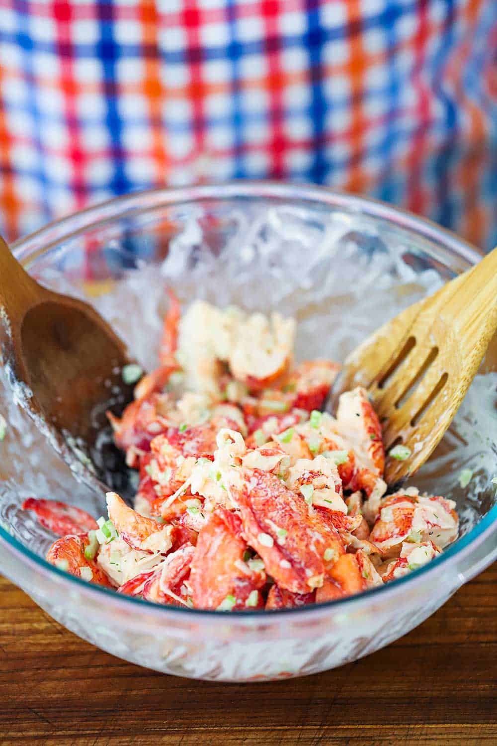 A glass bowl filled with lobster salad that is being tossed by a couple of wooden spoons. 