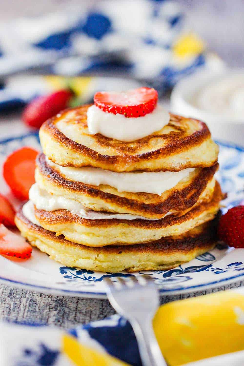 A stack of lemon ricotta pancakes with vanilla sauce on a antique plate with a fork nearby and strawberries. 