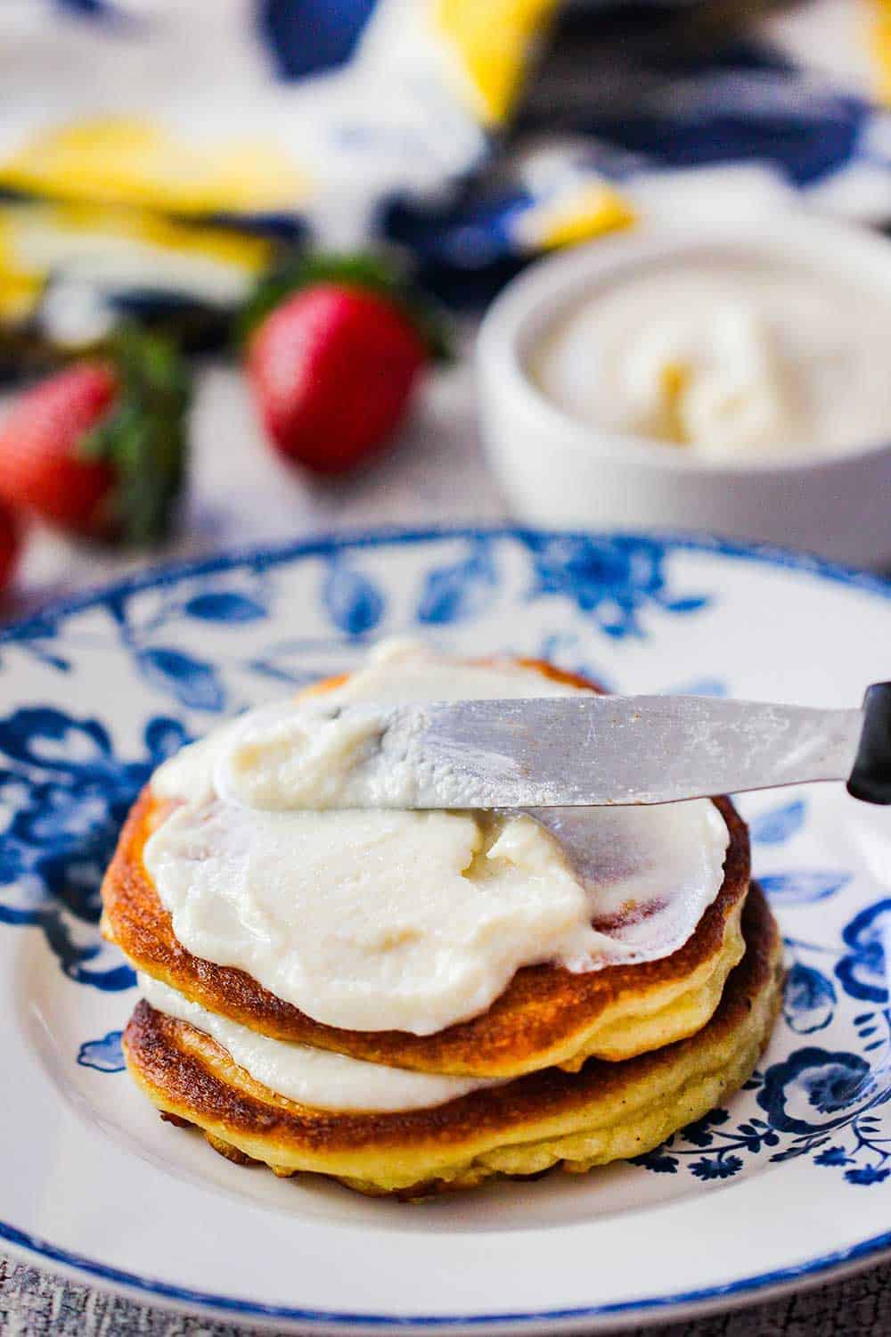 A knife smearing vanilla sauce onto lemon ricotta pancakes on a decorative plate. 