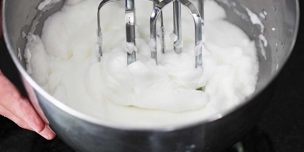 A close-up view of a hand mixer making stiff peaks from egg whites in a metal bowl. 