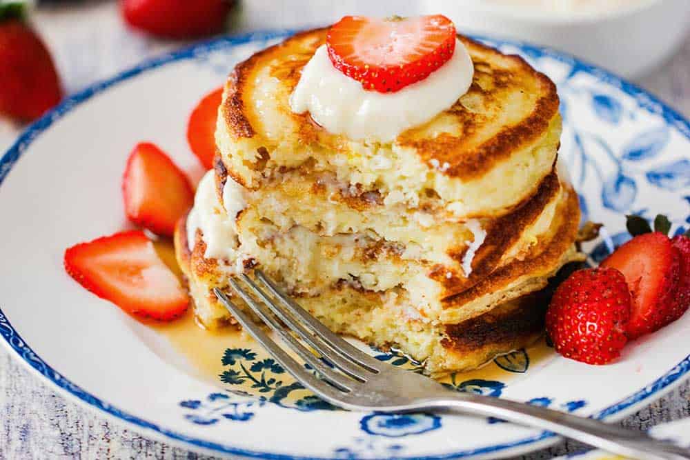 A stack of lemon ricotta pancakes that have been half eaten, with a fork nearby and strawberries on the plate, too. 