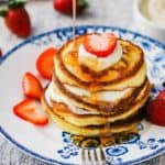 A stack of lemon ricotta pancakes with syrup being drizzled on top with strawberries nearby.