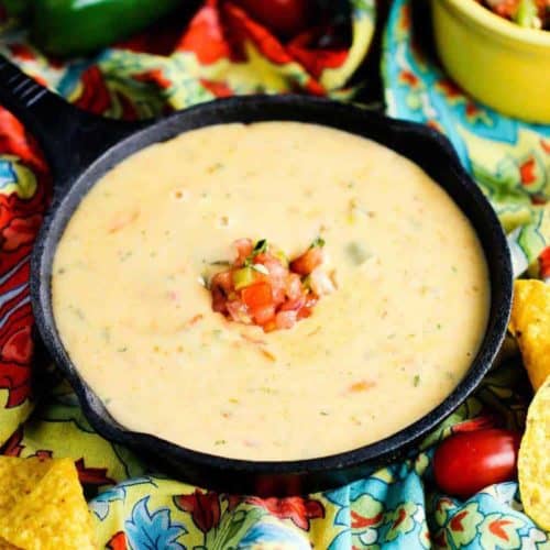 A bowl of Mexican cheese dip in a small cast iron skillet surround by peppers and festive napkins.