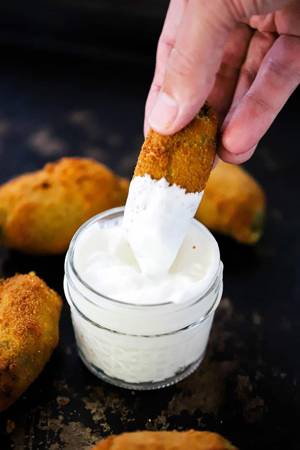 A hand dipping a breaded jalapeño into a small jar of sour cream. 