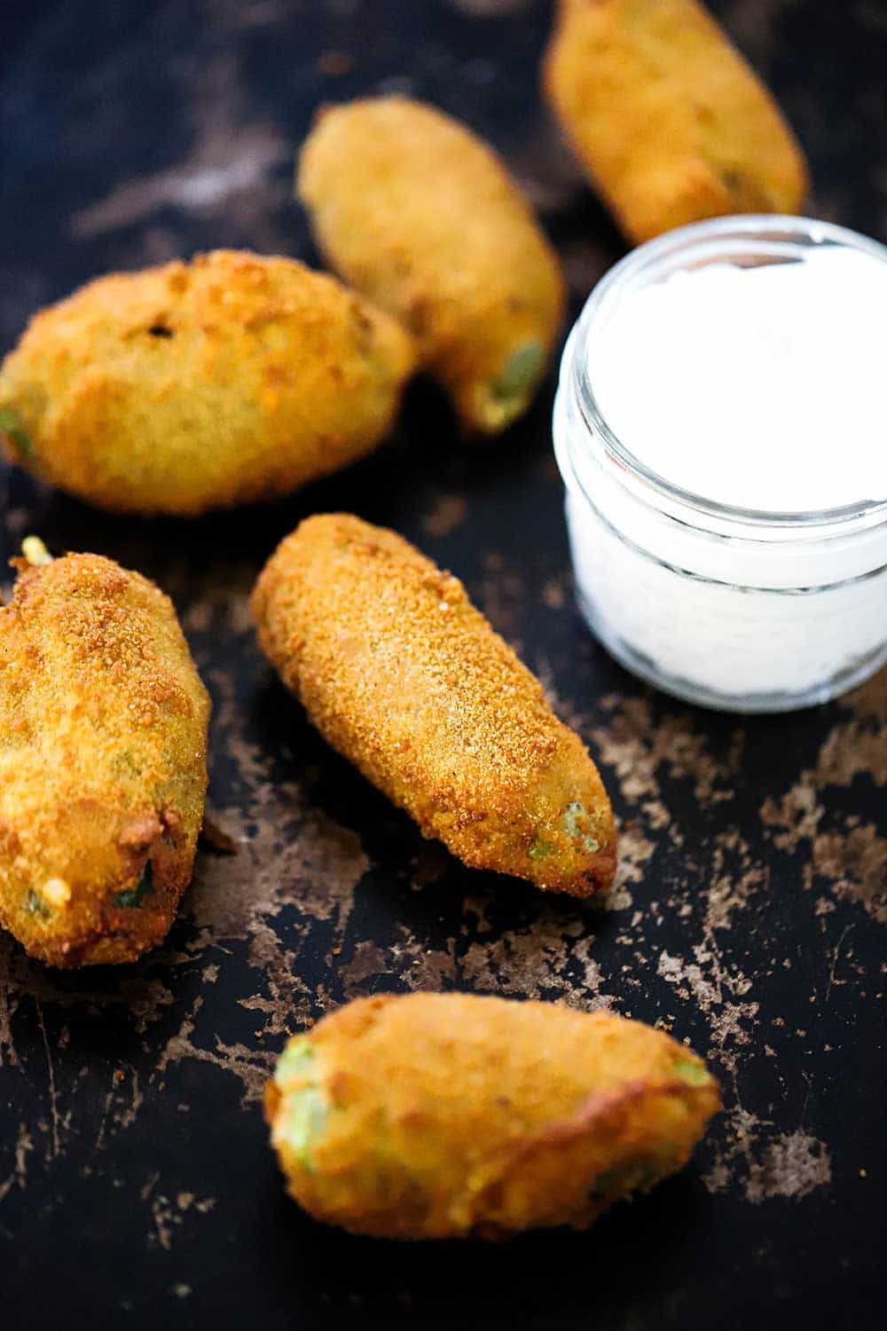 5 jalapeño poppers on a scratchy baking sheet next to a small jar of sour cream. 