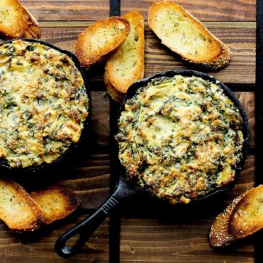 Two small cast iron skillet of baked spinach and artichoke dip with toasted bread nearby.