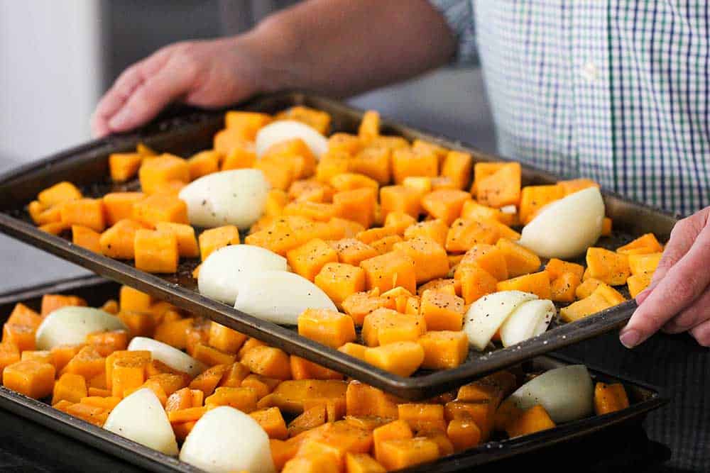 Two hands holding a baking sheet of cut butternut squash and onion . 