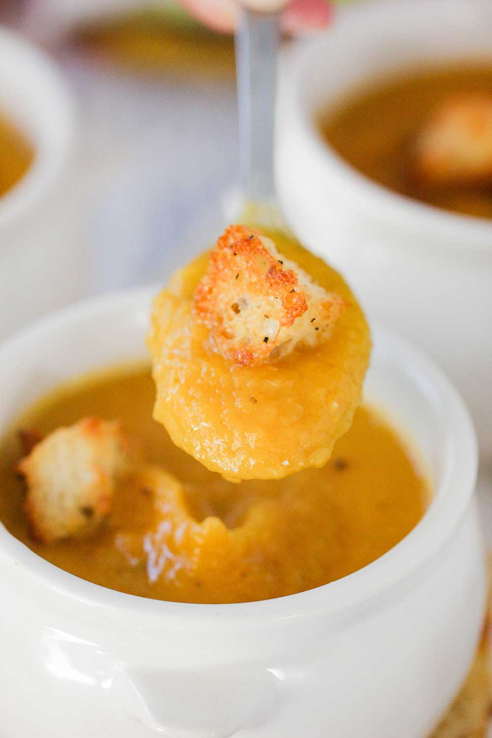 A hand holding a spoon full of roasted butternut squash soup over a small crock of soup with croutons on it. 