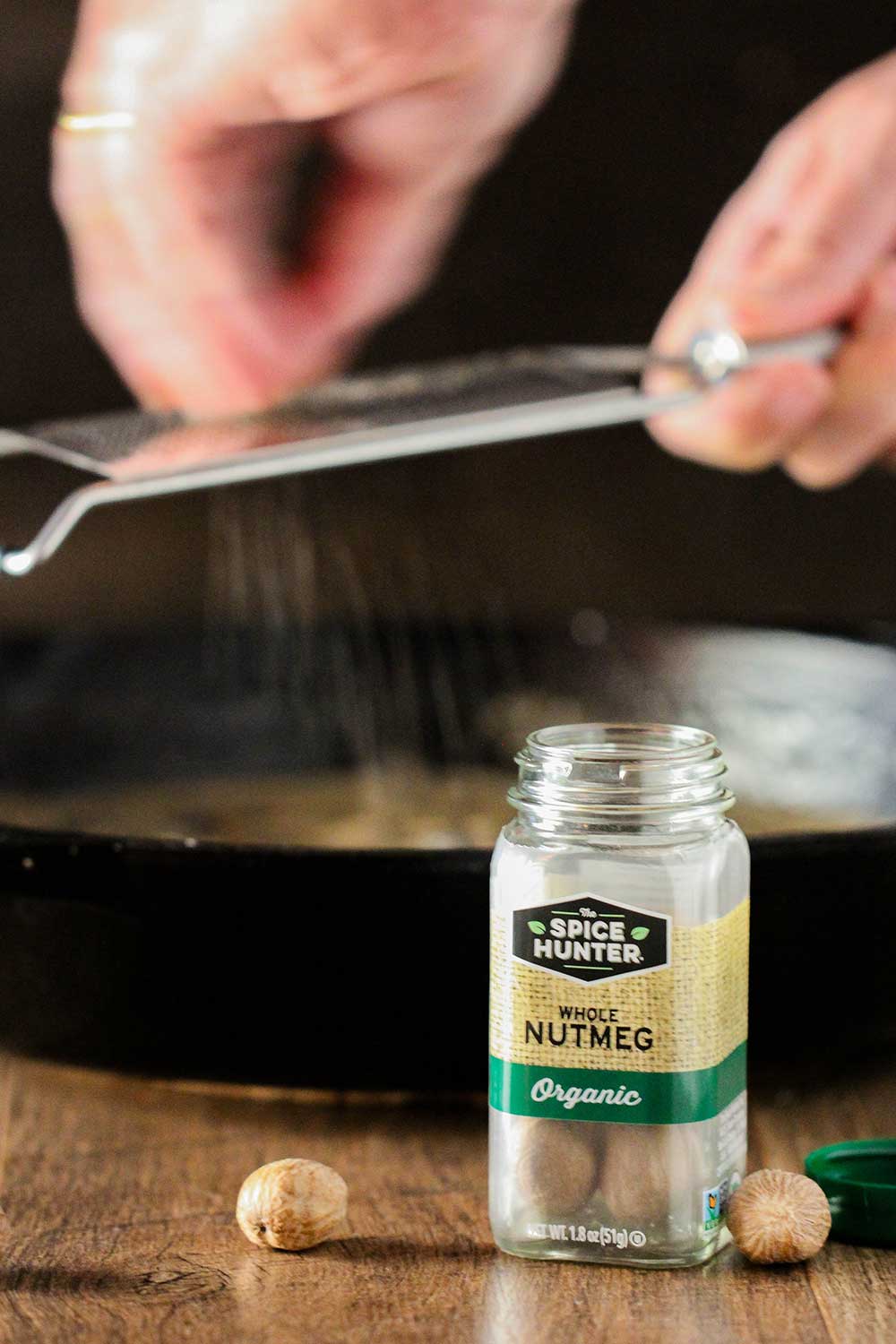 A hand scraping a whole nutmeg over a pan with a bottle of whole nutmegs in front of it. 