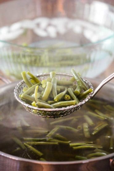 A slotted spoon holding cooked green beans over a skillet with a large bowl of ice water next to it.