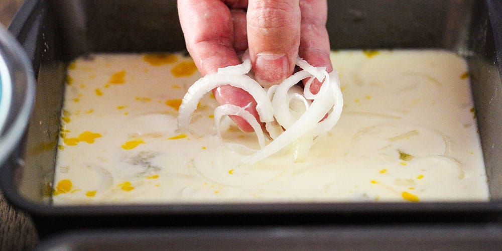 A hand dipping onion slices into a milk and egg bath in a metal pan. 