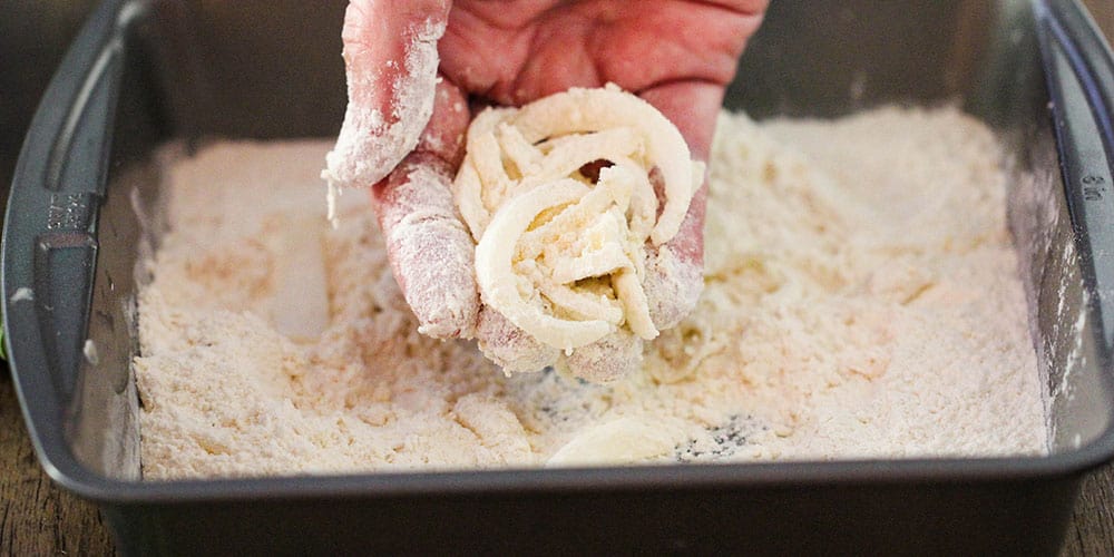A hand holding onion slices that have been dipped in milk and coated with flour in a metal pan. 