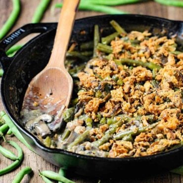 A large cast iron skillet filled with gourmet green bean casserole with a wooden spoon in it.