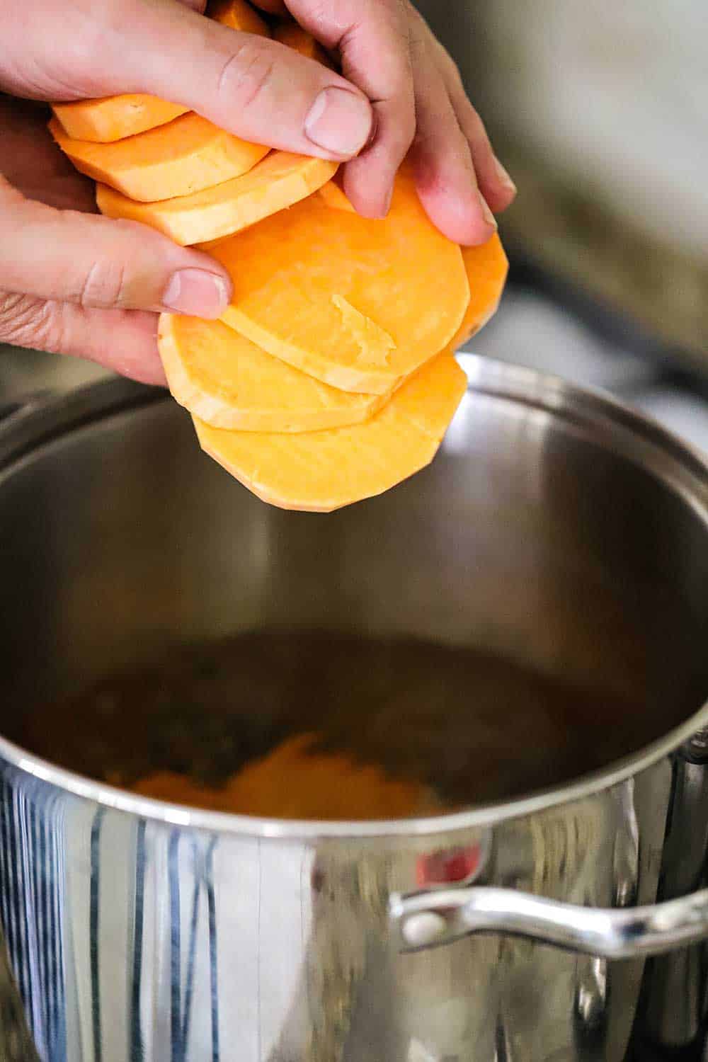 Two hands dropping sliced sweet potatoes into a pot of boiling water. 