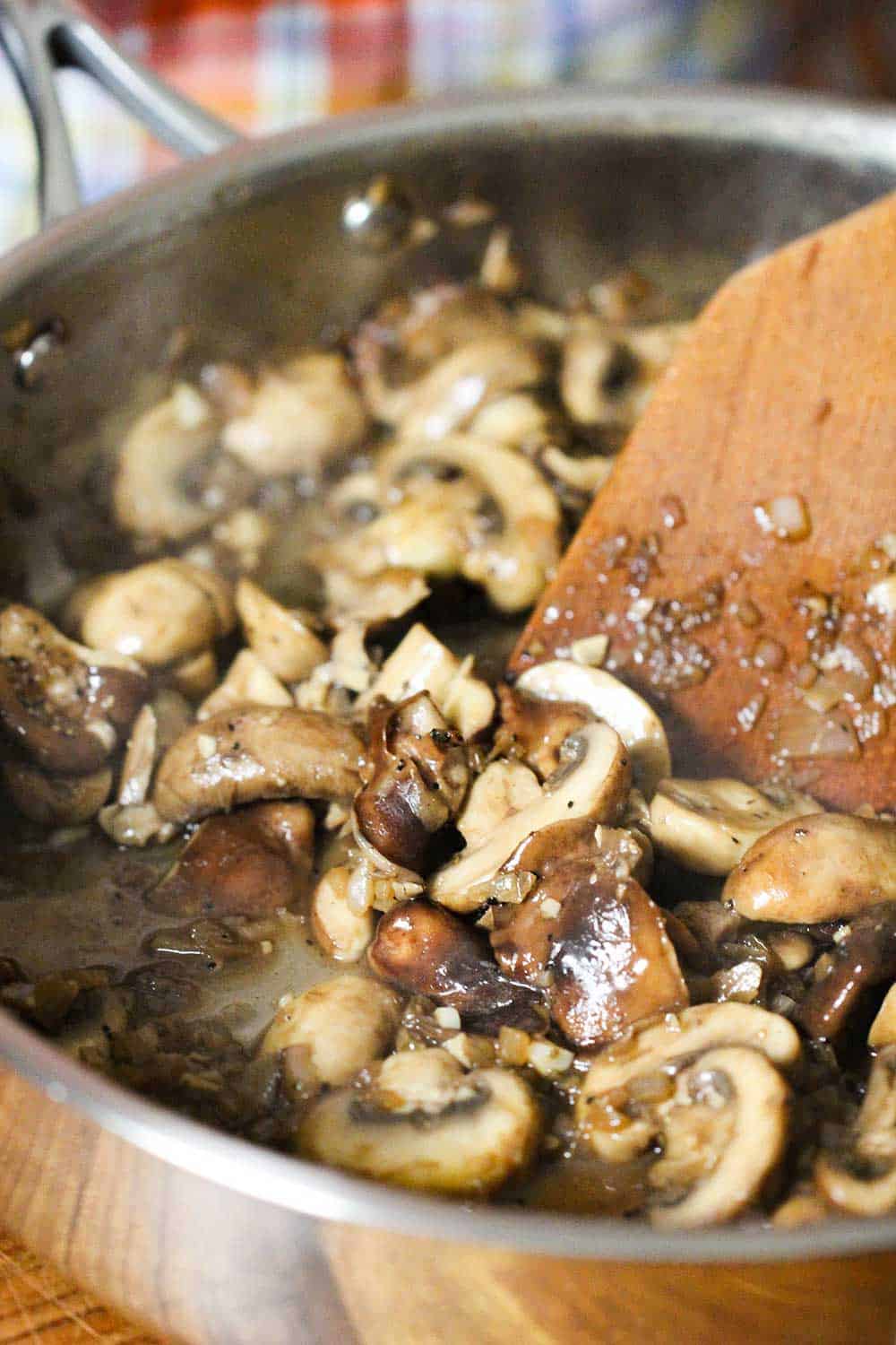 A large skillet with sautéed mushrooms in it being stirred by a wooden spoon. 