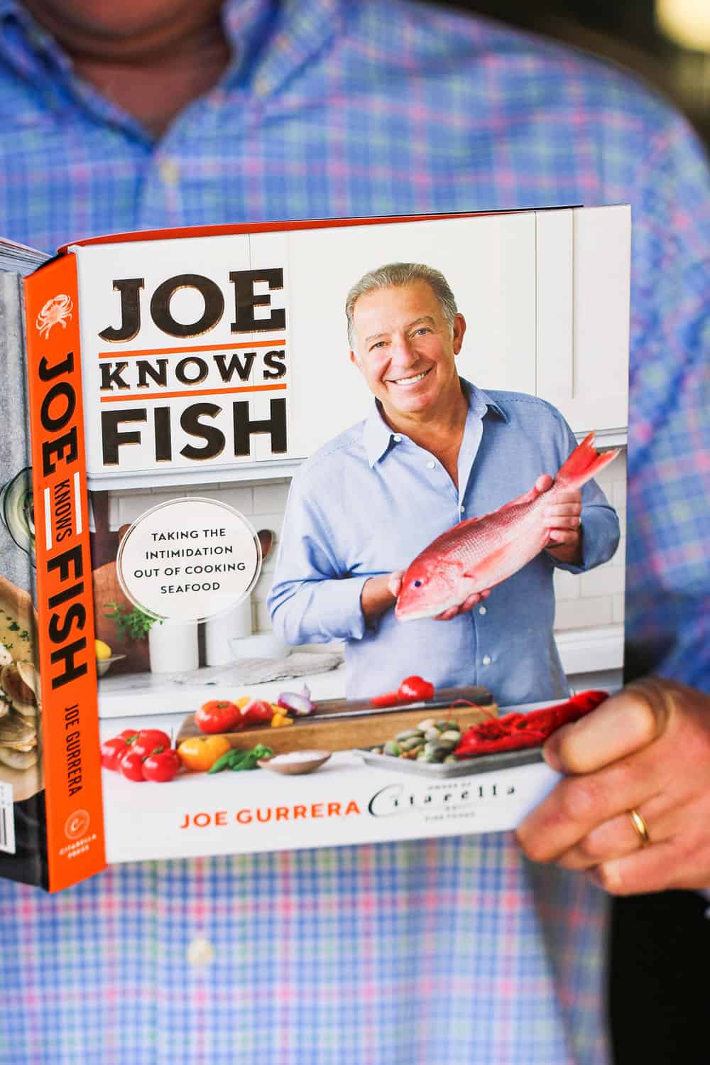 A male in a blue shirt hold the cookbook "Joe Knows Fish."