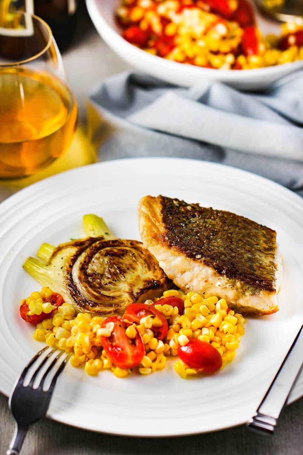 A white plate of sautéed wild striped bass with corn tomato salad and fennel