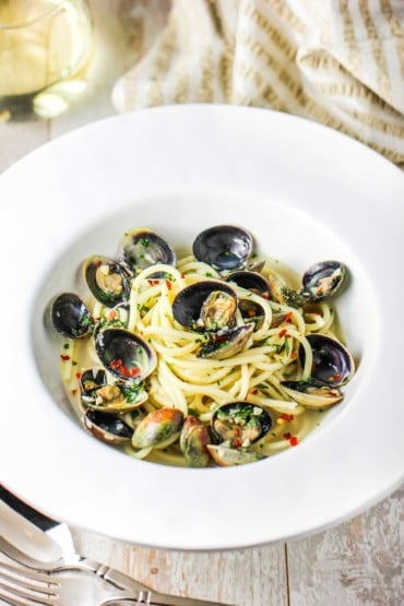 A large white bowl filled with spaghetti vongoles and a glass of white wine nearby.