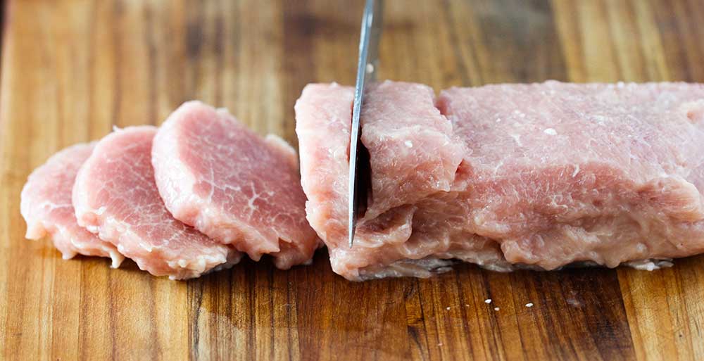 A knife slicing through an uncooked pork tenderloin on a cutting board. 