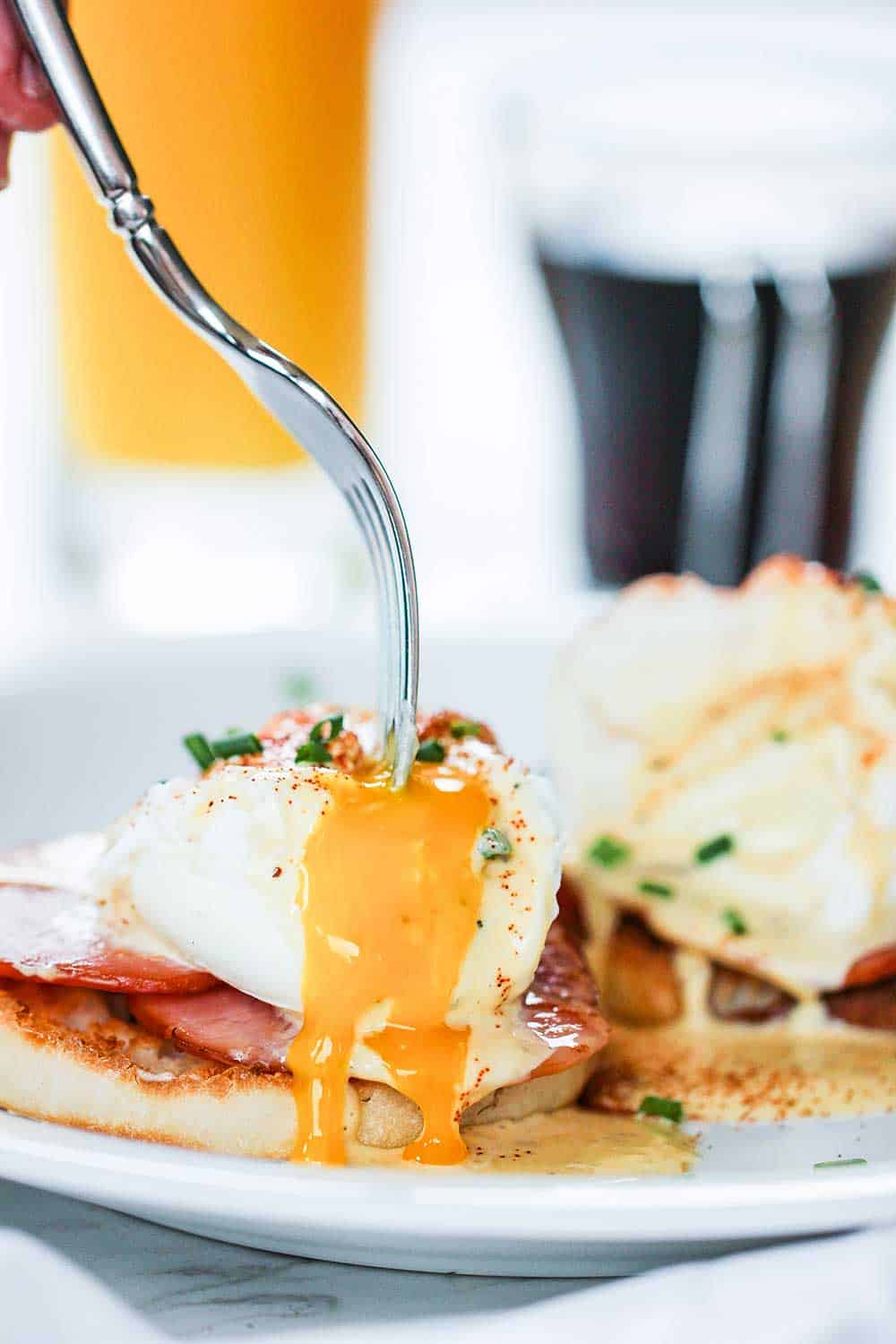 A fork puncturing a poached egg with yolk running on to Canadian bacon and an English muffin. 