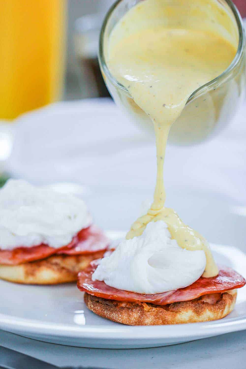 A small glass pitcher of Hollandaise sauce being poured onto eggs Benedict.