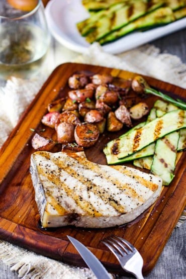 Grilled swordfish steak on a cutting board next to potatoes and grilled zucchini.