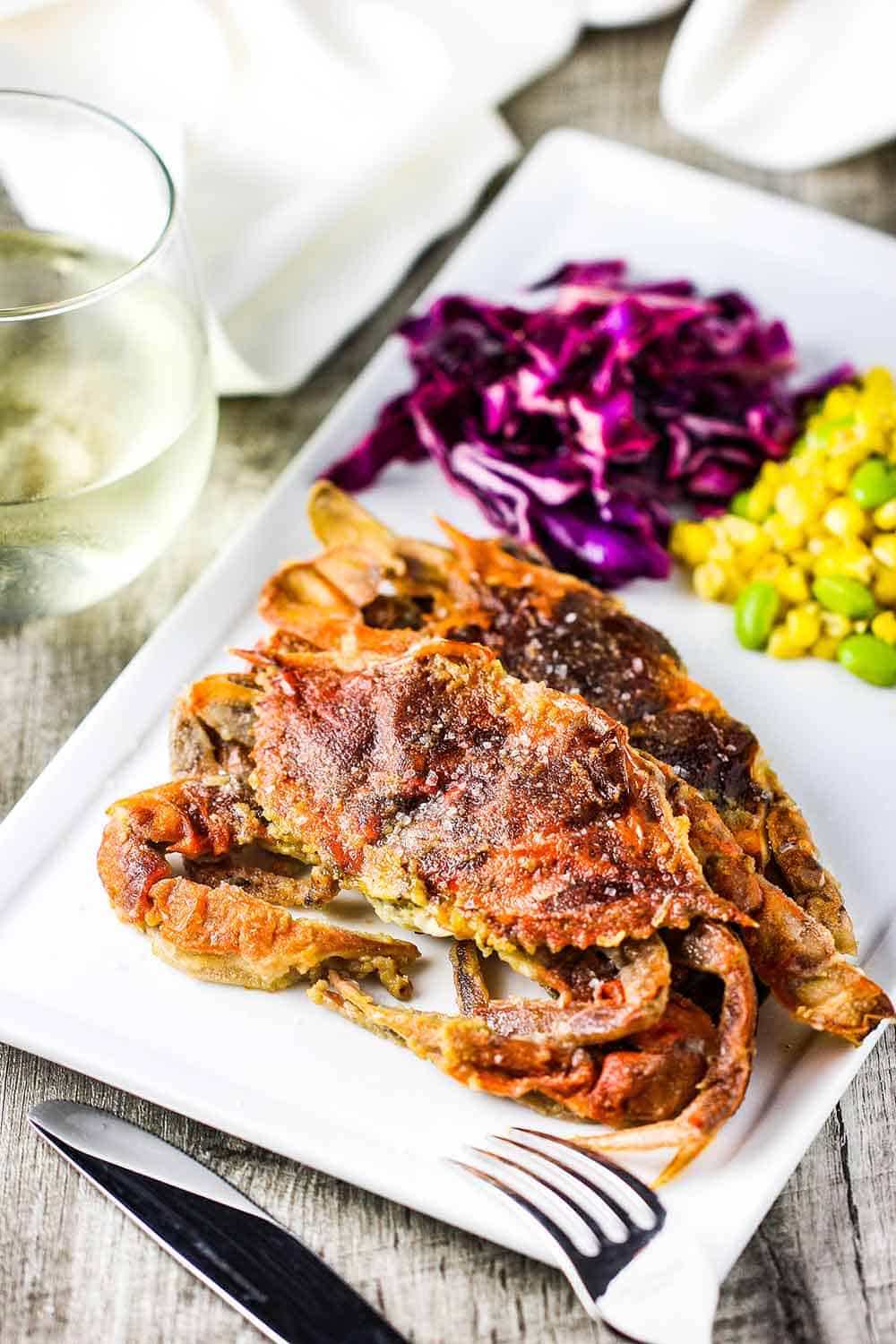 Two sautéed soft-shell crabs on a white plate with red cabbage and corn salad next to it. 