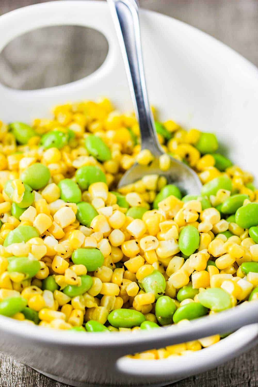A white bowl of fresh corn and edamame salad with a sliver spoon. 