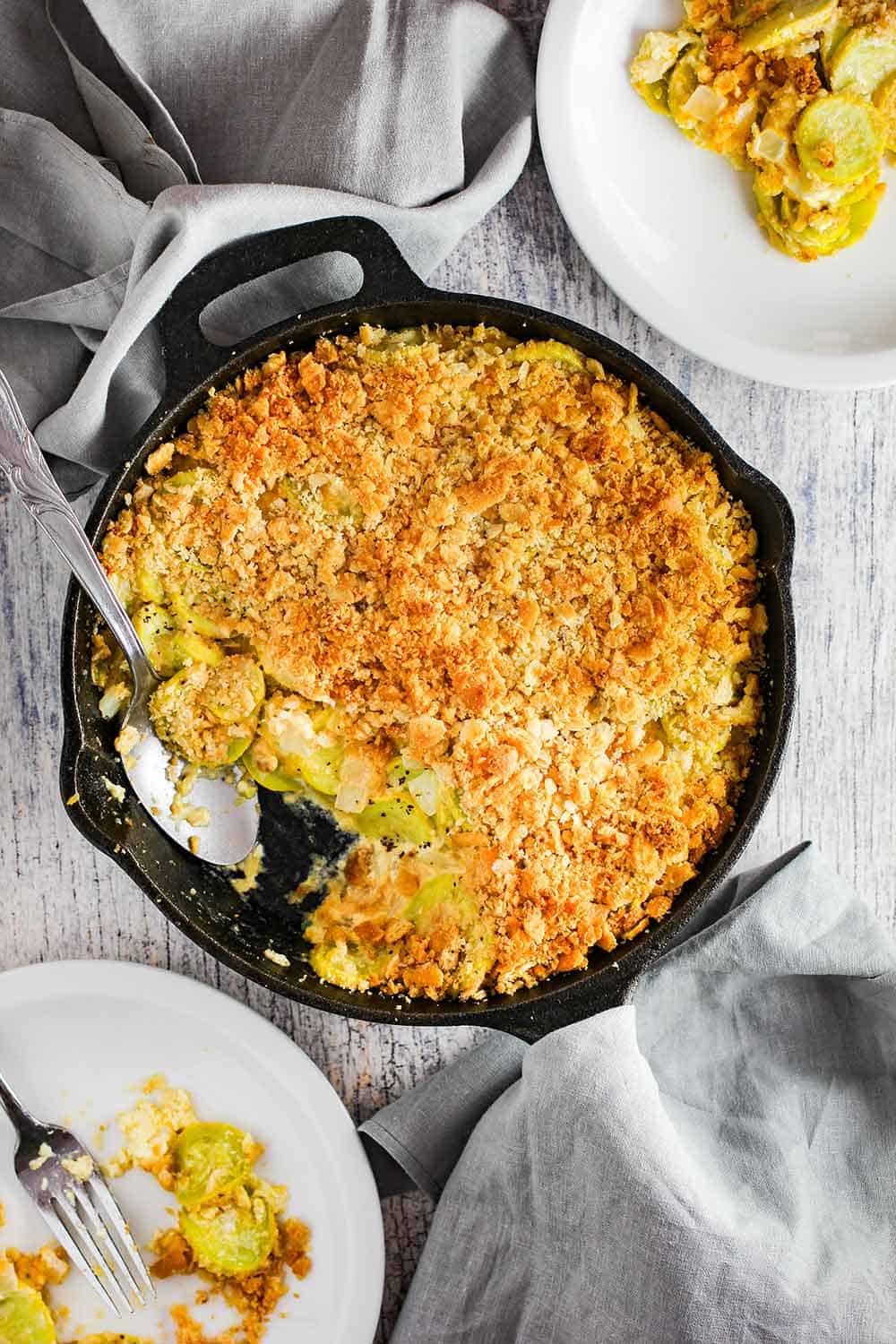 An overhead view of a cooked yellow squash casserole with two plates of food nearby. 