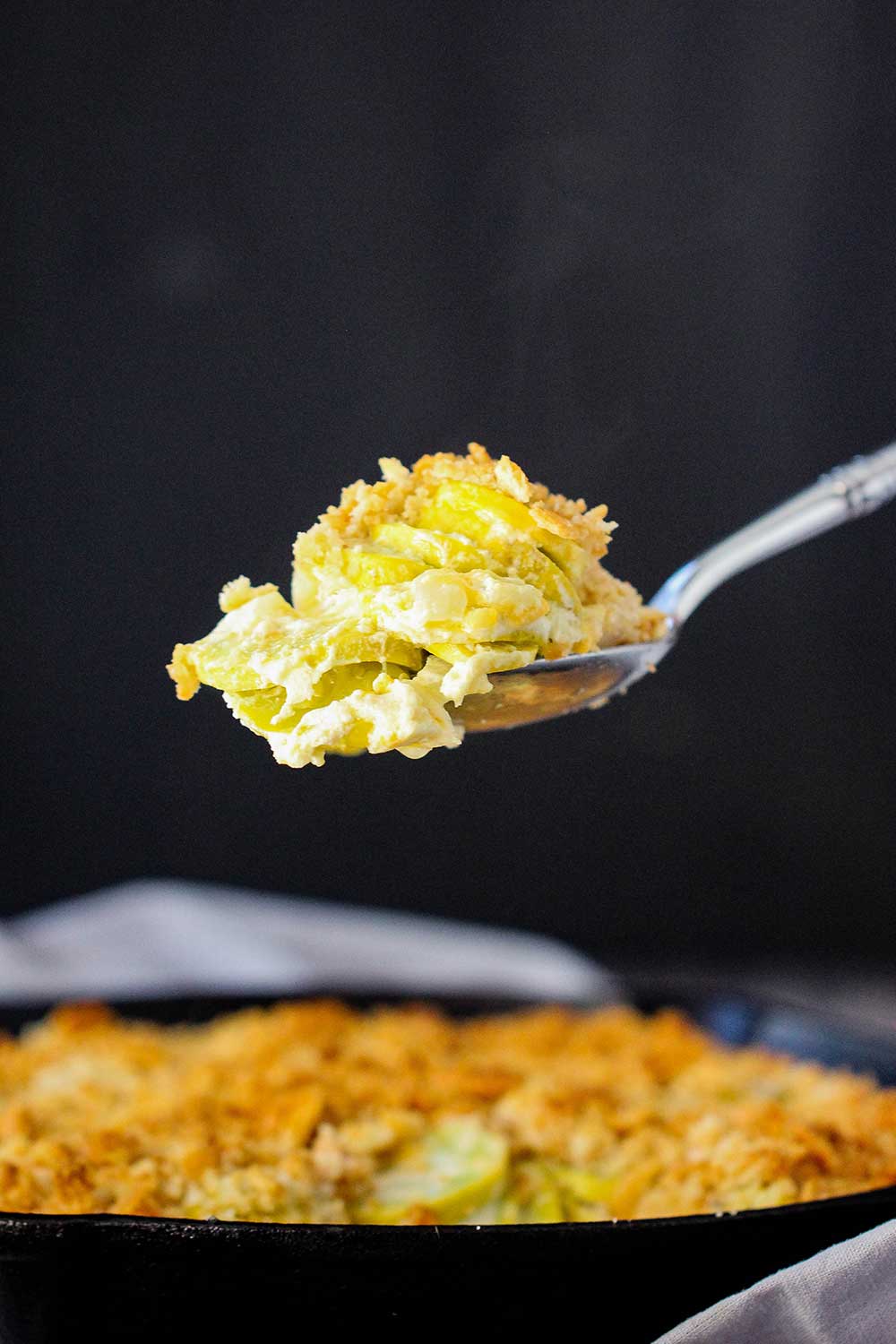 A spoon lifting up a large helping of cooked yellow squash casserole out of the skillet. 