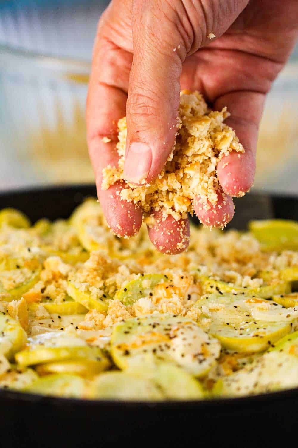 Fingers sprinkling crushed Ritz crackers over the casserole in a large black cast iron skillet. 