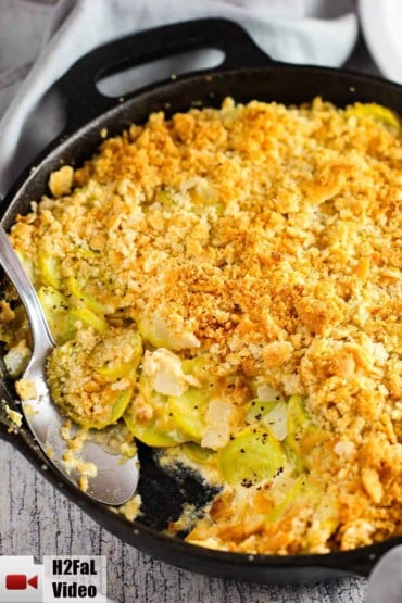 A large black cast iron skillet of yellow squash casserole with a serving spoon inserted in the dish.