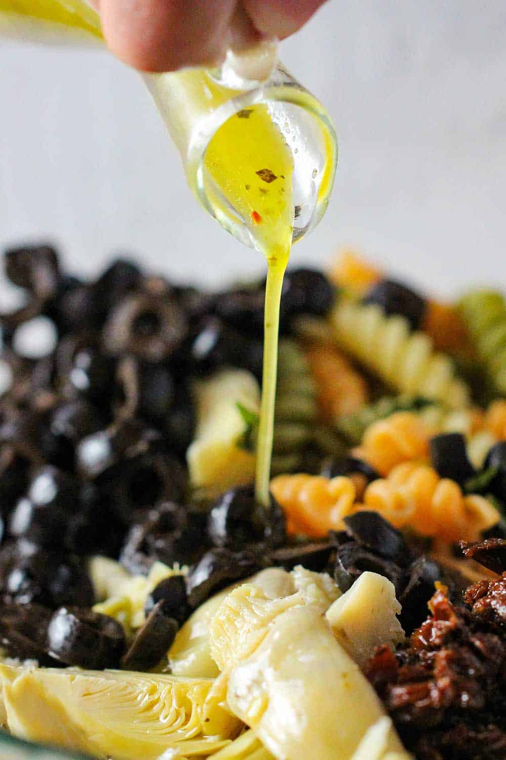 Italian dressing being pour onto a pasta salad with artichokes and black olives. 