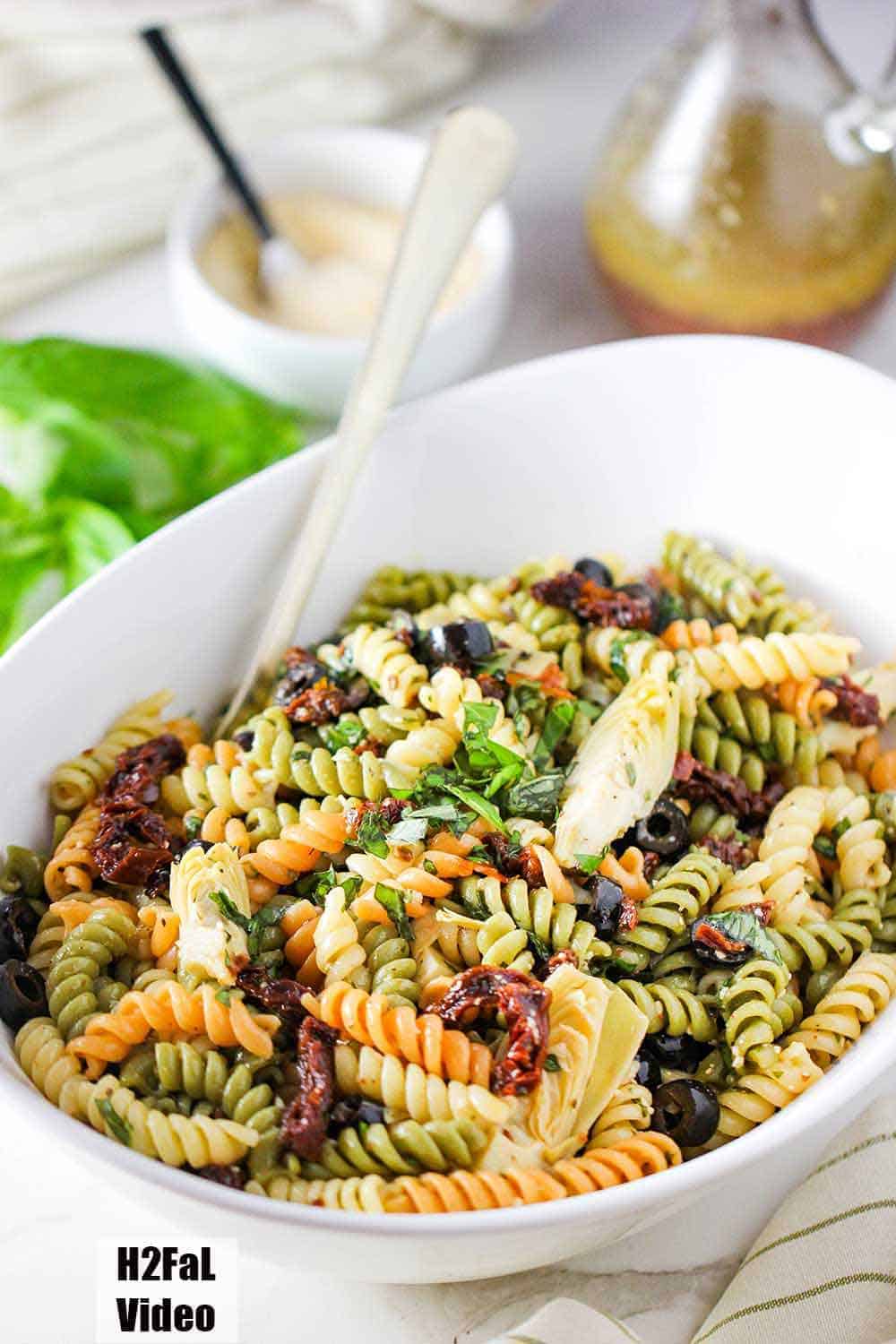 Classic Pasta Salad in a large white serving bowl with dressing and cheese nearby. 