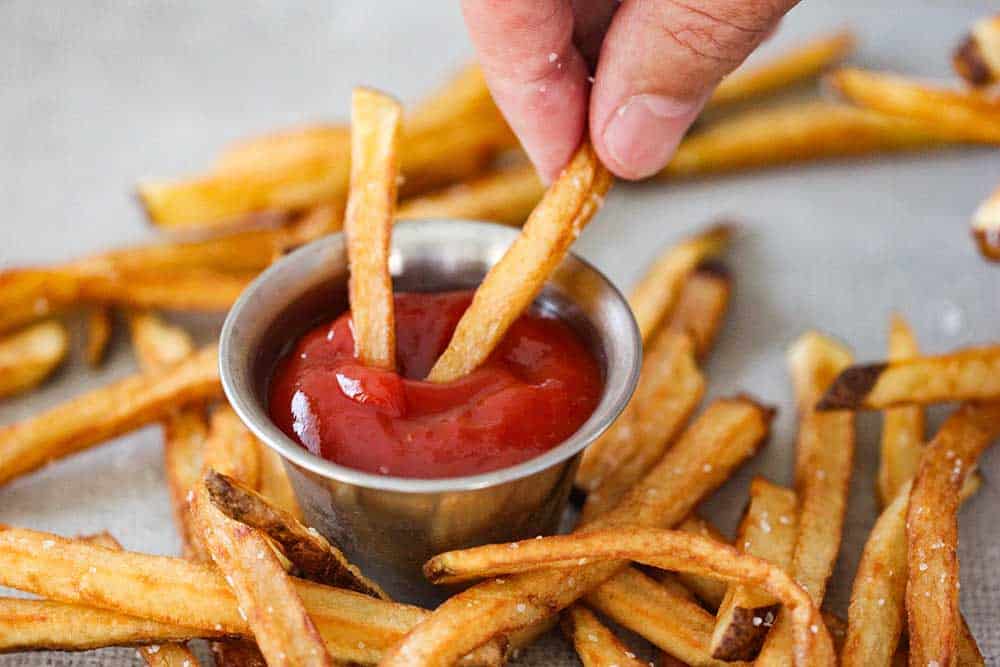  A hand dipping homemade French fries into a dish of ketchup. 