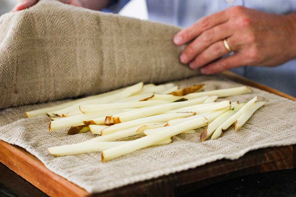 Two hands pressing a towel over cut potatoes for homemade French fries. 