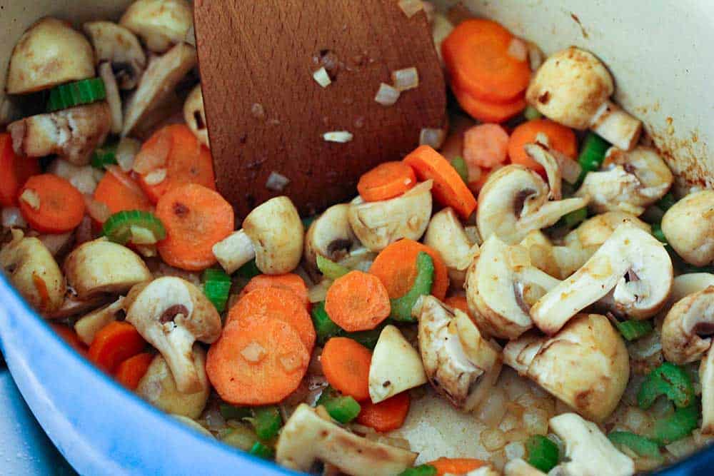 A wooden spoon moving around sautéed vegetables and mushrooms in a blue Dutch oven. 