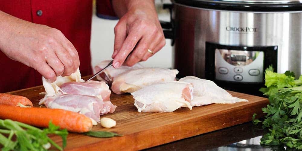 Two hands using a knife to remove skin from chicken thighs in front of a Crock-Pot. 