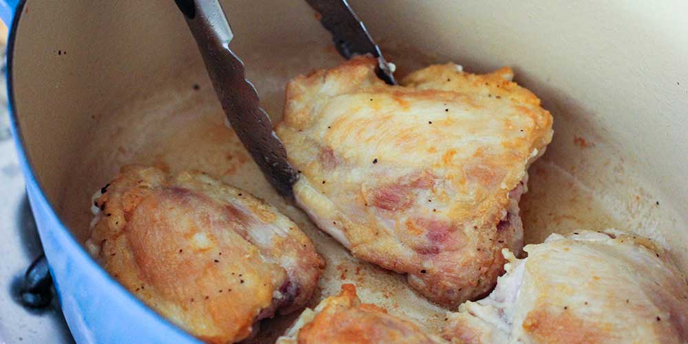 A pair of tongs flipping a seared chicken thigh in a large Dutch oven. 