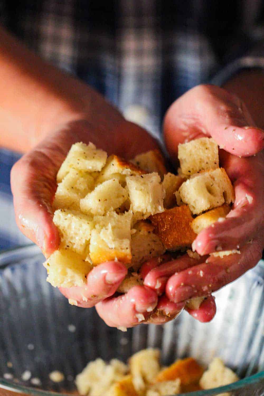 Two hands holding a mound of seasoned and oiled bread cubes over a glass bowl. 