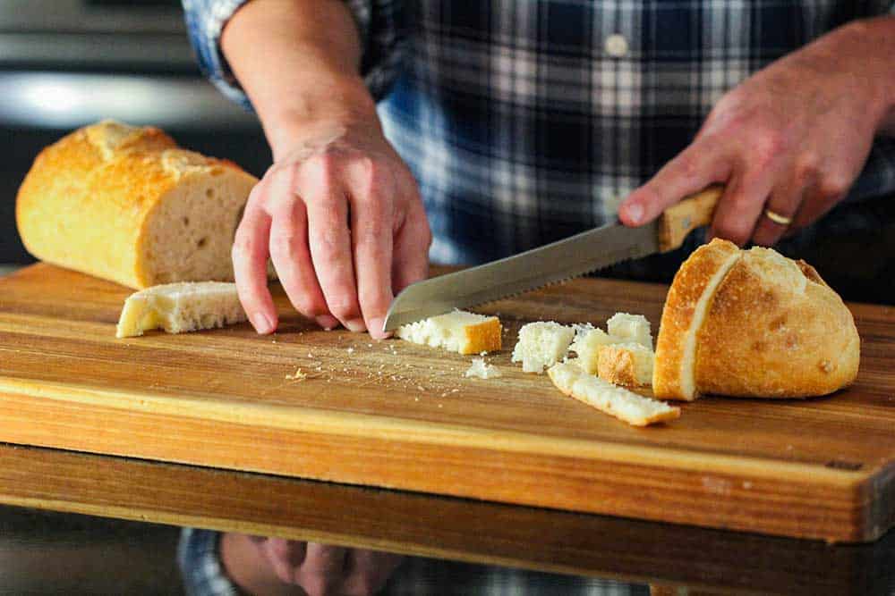 Two hands using a serrated knife to cut strips of bread on a cutting board. 