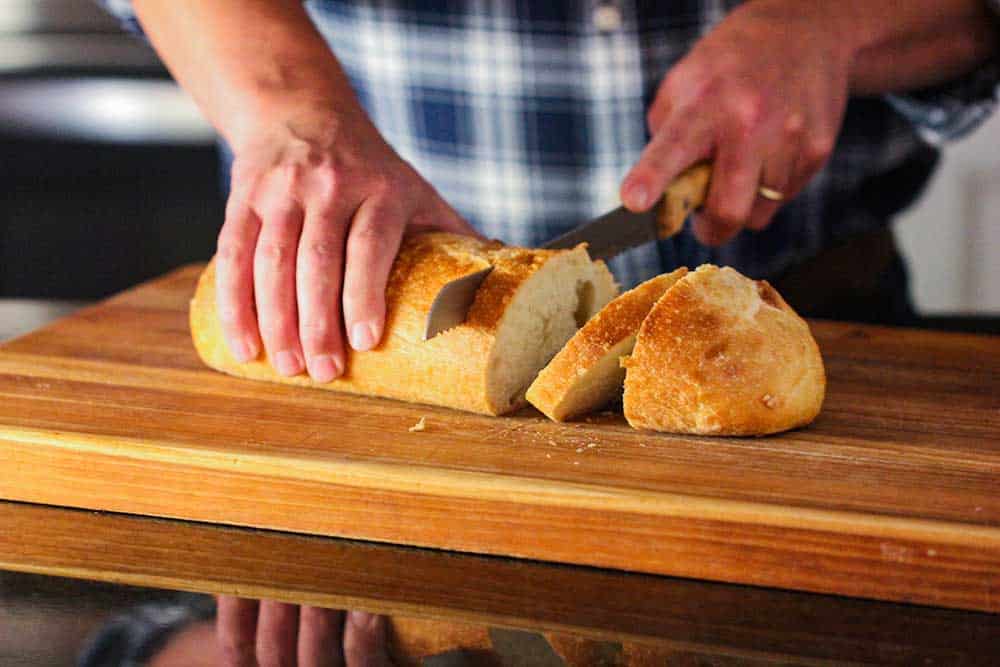 Two hands slicing through a loaf of Italian bread on a large wooden cutting board. 